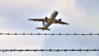Flugzeug startet am Flughafen, davor ein Zaun aus Stacheldraht.