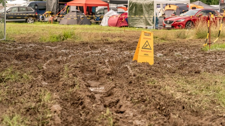 Mitten in einer matschigen Wiese auf dem Campinggelände der Nature One 2024 steht ein Warnschild mit der Aufschrift "Achtung, Rutschgefahr". 