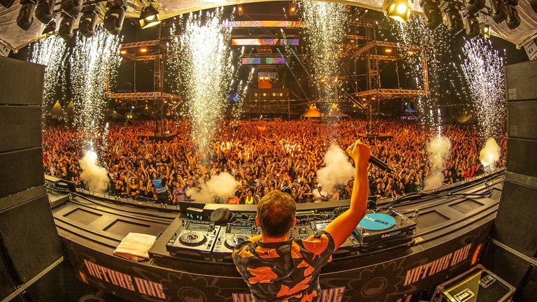 Ein DJ reckt auf der Bühne der NATURE ONE vor einer großen Menschenmenge sein Mikrofon in die Luft. Rund 65.000 Rave-Begeisterte werden dieses Jahr im Hunsrück erwartet.