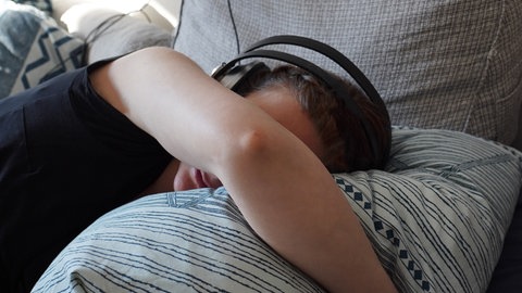 Eine Frau liegt im Bett mit Kopfhörern gegen Geräusche. Menschen, die an den schweren Folgen einer Corona-Infektion leiden, sind geräuschempfindlich und oft isoliert. 
