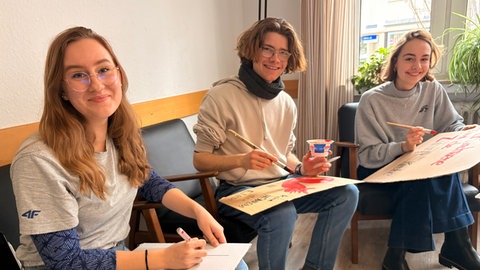 Drei junge Erwachsene sitzen zusammen und basteln Plakate mit roter Stift. Die Pappschilder tragen Parolen wie "ESGeht weiter!"