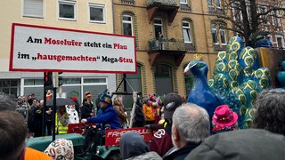 "Am Moselufer steht ein Pfau im hausgemachten Mega-Stau" steht auf einem großen Schild, ein Riesnpfau folgt dahinter.