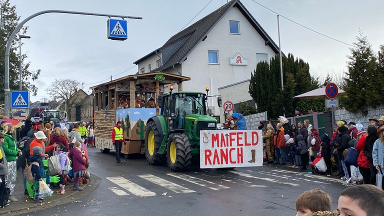 Ein Traktor zieht beim Rosenmontagsumzug 2026 in Münstermaifeld einen Karnevalswagen. Vorne hat er ein Schild mit der Aufschrift "Maifeld Ranch".
