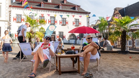Zwei Personen sitzen entspannt auf einem Liegestuhl auf dem Sandstrand in der Altstadt von Linz am Rhein. 