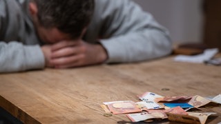 Ein Mann hat seinen Kopf auf seine Hände gelegt, während sich vor ihm Geldscheine und Münzen auf einem Tisch befinden: Viele Menschen in Ludwigshafen haben Geldsorgen. Jeder siebte Ludwigshafener hat Schulden.