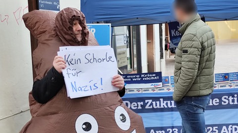 Mann protestiert in Neustadt in einem Kostüm, das wie ein Kackhaufen aussieht
