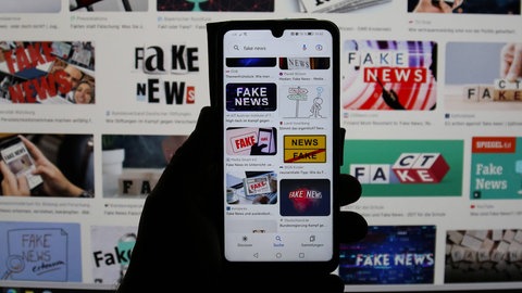 Hand hält Handy mit verschiedenen Schriftzügen "Fake News", Symbild für Artikel Fakenews im Zusammenhang mit dem Ludwigshafener, der in Mannheim in einem Menschenmenge gerast ist