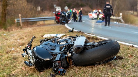 Zerstörtes Motorrad liegt am Straßenrand, im Hintergrund unscharf Rettungskräfte, Symbolbild für LKA und Polizei warnen vor Motorradunfällen zum Start der Motorradsaison