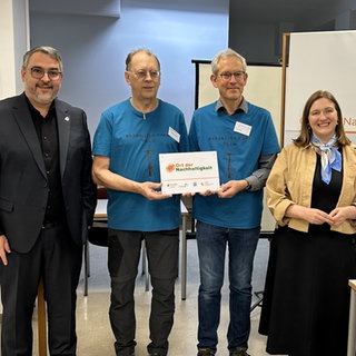 Neustadts Oberbürgermeister Marc Weigel, zwei Mitarbeitende des Reparatur-Cafés in Neustadt und Verbraucherschutzministerin Katharina Binz (v.l.n.r.). Die Ehrenamtler halten eine Auszeichnung in der Hand. Das Café wurde zum Ort der Nachhaltigkeit ernannt.