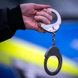 Ein Polizist vor einem Polizeiauto hält ein Paar Handschellen (Symbolbild): In Wörth am Rhein ist ein Mann nach einer Schlägerei im Bahnhof festgenommen worden.