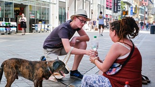 Ein Mann reicht einer obdachlosen Frau auf dem Boden ein Wasser. Symbolbild. Was machen Obdachlose in der Pfalz bei großer Hitze?