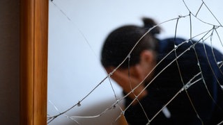 Eine Frau sitzt neben einem gesprungenen Spiegel auf einem Bett und hält ihren Kopf in den Händen (gestellte Szene)
