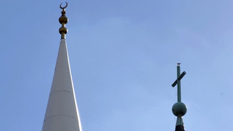 Eine Sichel auf dem Minarett einer Moschee und ein Kreuz auf einem Kirchturm. 