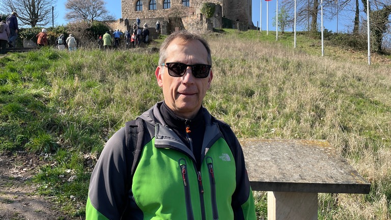 Michael Gans aus Neustadt-Mußbach steht auf einer Treppe, die zum Hambacher Schloss führt. Er war mit einer Seniorengruppe unterwegs und machte mit ihr eine Wanderung zum Hambacher Schloss. Er wollte den schönen Ausblick von oben genießen und einfach in Ruhe einen Kaffee trinken. Er freut sich, dass das Hambacher Schloss sich am Aktionstag beteiligt. Er findet es positiv, dass es den Tag der Demokratiegeschichte gibt und "dass man sich die Demokratie vor Augen hält".