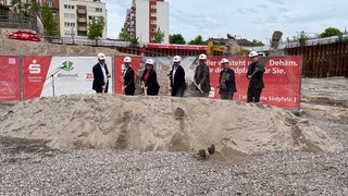 Hier wird geschaufelt: Sieben Menschen beim offizielle Spatenstich für das neue Sparkassengebäude in Landau, darunter der Landrat des Kreises Südliche Weinstraße Dietmar Seefeldt (ganz links) und der Oberbürgermeister der Stadt Landau Dominik Geißler (ganz rechts).