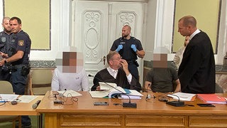 die beiden Verurteilten Mitglieder einer Schockanruferbande im Gerichtssaal des Landgerichts Landau mit ihren Anwälten. Im Hintergrund Justizbeamte.