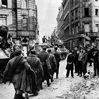 Eine Gruppe Jugendlicher steht am Straßenrand (r) und beobachtet den Einmarsch von US-Soldaten und Panzern in Mainz gegen Ende des Zweiten Weltkriegs.