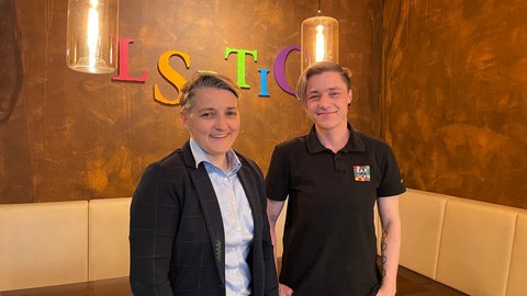 Queeres Zentrum: In der "Bar jeder Sicht" in Mainz stehen Diana Gläßer und Chey Schulmerich nebeneinander vor einem LSBITQ-Schriftzug.