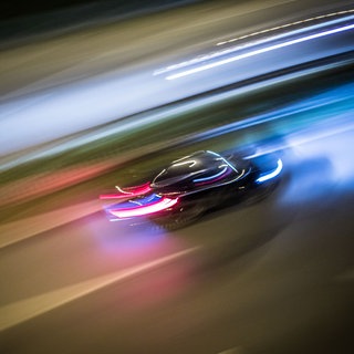 Ein Auto fährt sehr schnell auf einer Straße, die Scheinwerfer sind zu Lichtspuren verwischt.