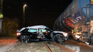 Das zerstörte Auto steht nachts quer auf einem Bahngleis, daneben steht ein Güterzug.