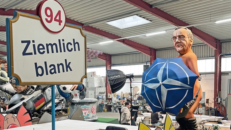 Verteidigungsminister Boris Pistorius (SPD) steht nackt auf dem Fastnachtswagen. Sein Geschlecht verdeckt er mit einem Schirm, auf dem das NATO-Logo abgedruckt ist.