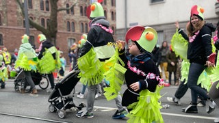 Ein kleiner grüner Halsbandsittich steht auf der Straße und bläst eine Luftschlange auf - beim 67- Kinder- und Jugendmaskenzug in Mainz. Er gilt als einer der größten Kinderumzüge Europas.