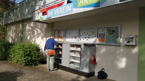 In einem draußen zugänglichen Regal liegen abgepackte Lebensmittel, ein Mann steht davor.