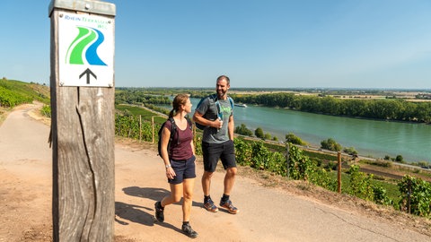 Eine Frau und ein Mann wandern auf dem Rheinterrassenweg oberhalb des Rheins. Der Wanderweg führt von Worms bis Mainz und ist an zwölf Haltestellen mit der Bahn erreichbar