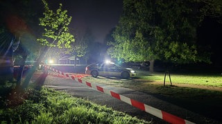 Ein Polizeiauto steht im dunklen Hartenbergpark. Der ist mit einem rot-weißen Flatterband abgesperrt.