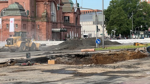 Im Straßenbelag ist ein Loch. Im Hintergrund sieht man die rote Fassade des Wiesbadener Hauptbahnhofs.