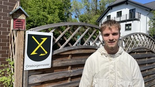 Der jüngste Ortsbürgermeister Deutschlands steht vor seinem Elternhaus in Eckenroth. Am Zaun hängt ein Schild mit dem Ortswappen und der Aufschrift Ortsbürgermeister.