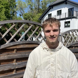 Der jüngste Ortsbürgermeister Deutschlands steht vor seinem Elternhaus in Eckenroth. Am Zaun hängt ein Schild mit dem Ortswappen und der Aufschrift Ortsbürgermeister.