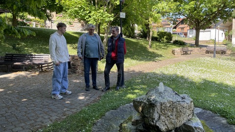Tim Sieper, sein Vater und der Erste Beigeordnete stehen auf dem grünen Dorfplatz von Eckenroth und sehen nach, ob an dem kleinen Brunnen alles in Ordnungs ist.