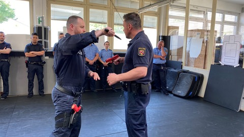 In einer Sporthalle stehen zwei Polizisten einander gegenüber, einer hat ein Messer gezückt, der andere eine Waffe. Am Rand stehen andere Polizisten und schauen zu.