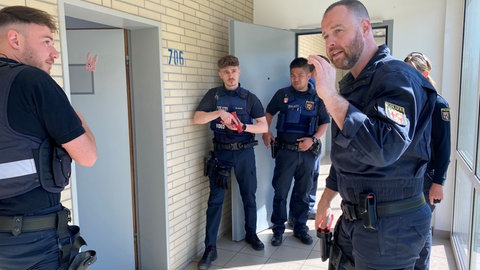 Mehrere Polizisten stehen vor einer geöffneten Tür, einer erklärt den anderen etwas. Ein anderer hat eine rote Waffe gezückt.
