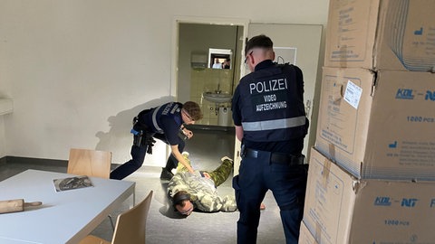 Ein Mann liegt bäuchlings auf dem Boden, ein Polizist hält seinen Arm fest. Ein anderer Polizist hat eine Waffe auf den Mann gerichtet. 