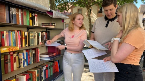 Drei Studentinnen der Buchwissenschaft füllen an einem Bücherschrank in Mainz-Mombach Listen aus, in denen sie den Inhalt des Schranks dokumentieren.