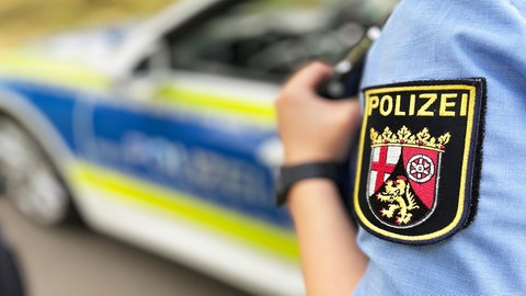 Ein Polizeiauto und eine Polizistin mit Wappen am Ärmel. In Mainz-Mombach haben sich zwei Familien auf dem Parkplatz eines Supermarktes geprügelt