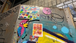 Auf einem stählernen Gerüst sind Aufkleber, eine bunte Fliese mit der Aufschrift "Glaube an dich selbst", ein Papagei und eine pinkfarbene Platte angebracht.