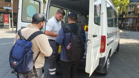 Jonathan Gaß, ehrenamtlicher Mitarbeiter des DRK Mainz-Hechtsheim, gibt zwei wohnungslosen Männern Wasser, Obst und Sonnencreme aus dem Hitzebus.