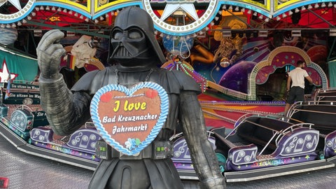 Eine Darth-Vader-Figur steht vor einem Fahrgeschäft auf der Pfingstwiese. Er trägt ein Lebkuchenherz mit der Aufschrift "I love Bad Kreuznacher Jahrmarkt". Damit ist er nicht alleine. Es werden bis zu 300.000 Besucherinnen und Besucher zu dem fünftägigen Volksfest erwartet 