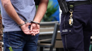 Ein Mann wird mit Handschellen von einem Polizisten abgeführt. (Symbolbild) In Worms soll ein Mann eine Seniorin getötet haben. 