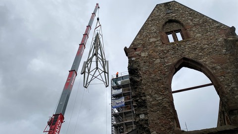 Eine Turmspitze aus Stahl wird an Seilen von einem Kran gehalten. Sie schwebt neben der Kirchenruine St. Christoph in Mainz in der Luft.