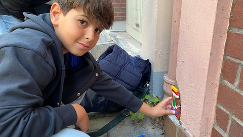 Elyas Bikri sitzt auf dem Bürgersteig. An der Hauswand vor ihm ist der Putz abgeplatzt. In das Loch passt Elyas sein Lego-Bauwerk ein.