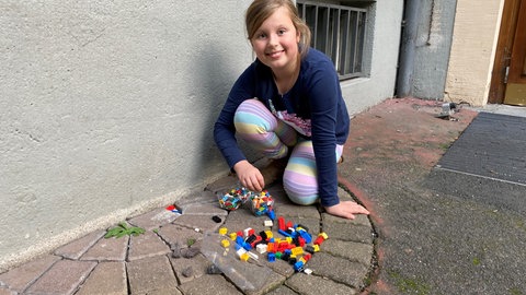 Die achtjährige Luise kniet auf dem Schulhof, vor ihr Legosteine, die sie in ein Loch auf dem Boden setzt.
