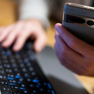 Ein Mann sitzt am Laptop und hält ein Smartphone mit in der Hand. In Mainz haben mehrere Menschen viel Geld an Anlagebetrüger verloren. 