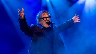 Sven Regener steht vor einem Mikrofon und reißt die Arme hoch: Der Schriftsteller und Musiker wird der Mainzer Stadtschreiber 2026