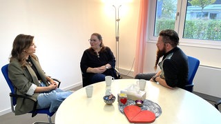 Zwei Frauen und ein Mann sitzen am Tisch, blicken sich an und unterhalten sich. In der Trauergruppe des Mainzer Hospiz werden junge Verwitwete unterstützt.