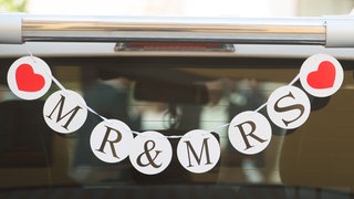 Eine Girlande mit den Buchstaben MR und MRS sowie zwei roten Herzen hängt an der Heckscheibe eines Autos.