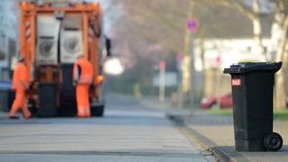 Eine Mülltonne steht auf der Straße, im Hintergrund ein Müllauto